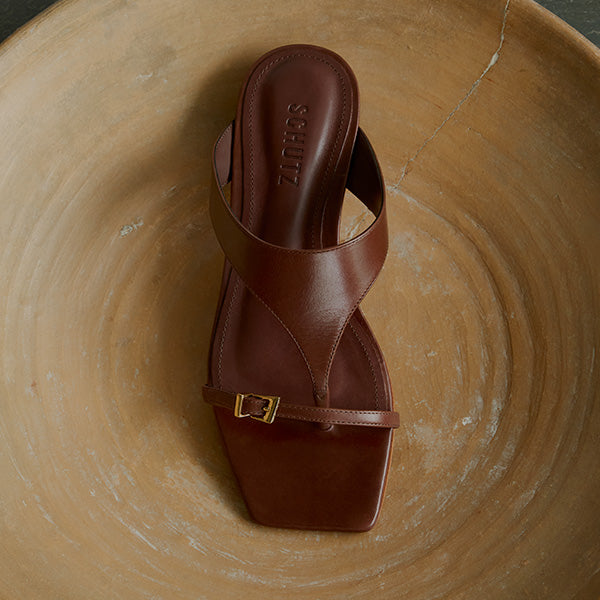 A single brown leather sandal with a square toe and thin strap, displayed on a large, round, tan ceramic dish.