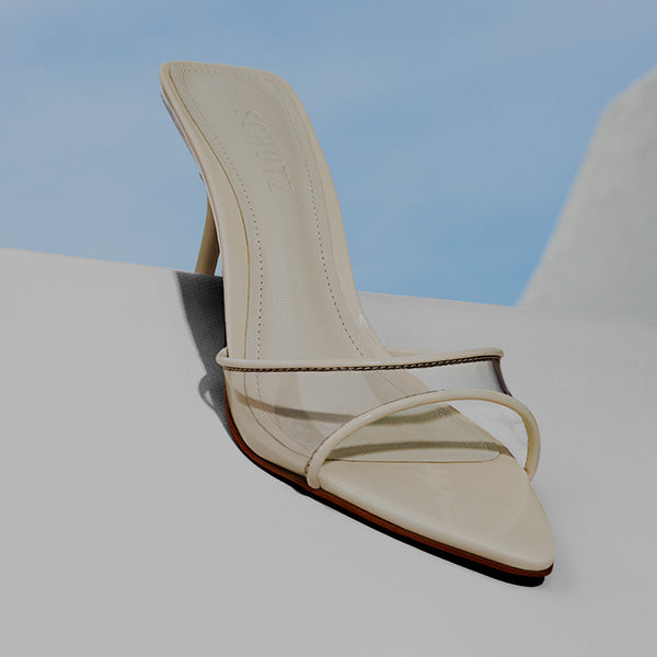 A beige heeled sandal with a pointed toe and transparent strap is displayed against a light background with a blue sky.