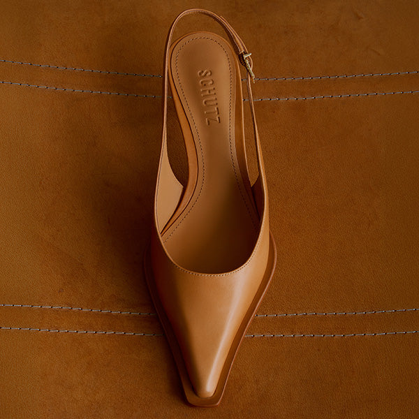 A tan leather pointed-toe slingback shoe on a matching tan leather surface, with stitched lines. Elegant and minimalist design conveys sophistication.