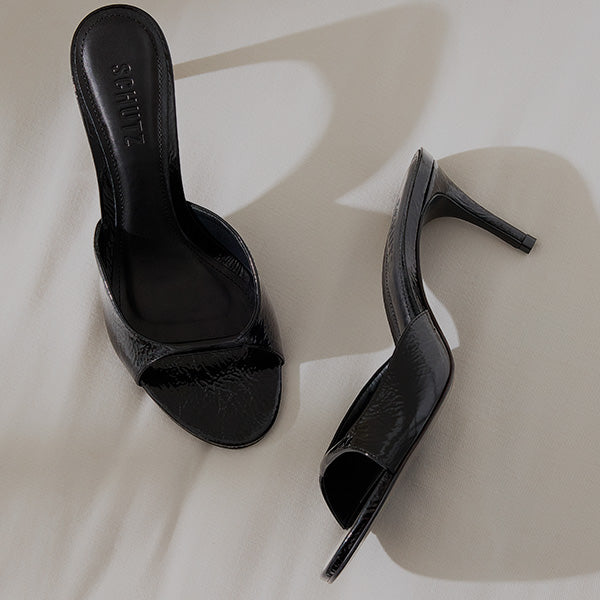 Black high-heeled mules with a glossy finish rest on a light surface, casting soft shadows. The open-toe design conveys elegance and style.