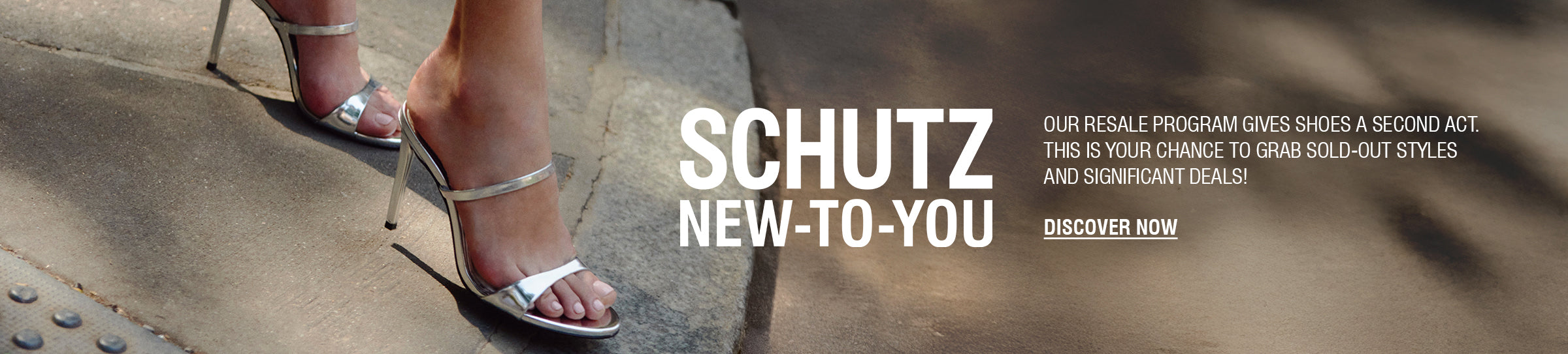 A person's legs and feet are visible on a crosswalk. They are wearing a pair of silver metallic high-heel mules with two thin straps over the foot. The text "SCHUTZ NEW-TO-YOU" and "DISCOVER NOW" are on the right side of the image, advertising a resale program.