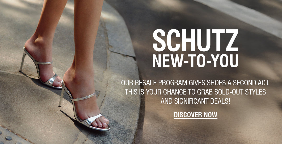 A person's legs and feet are visible on a crosswalk. They are wearing a pair of silver metallic high-heel mules with two thin straps over the foot. The text "SCHUTZ NEW-TO-YOU" and "DISCOVER NOW" are on the right side of the image, advertising a resale program.