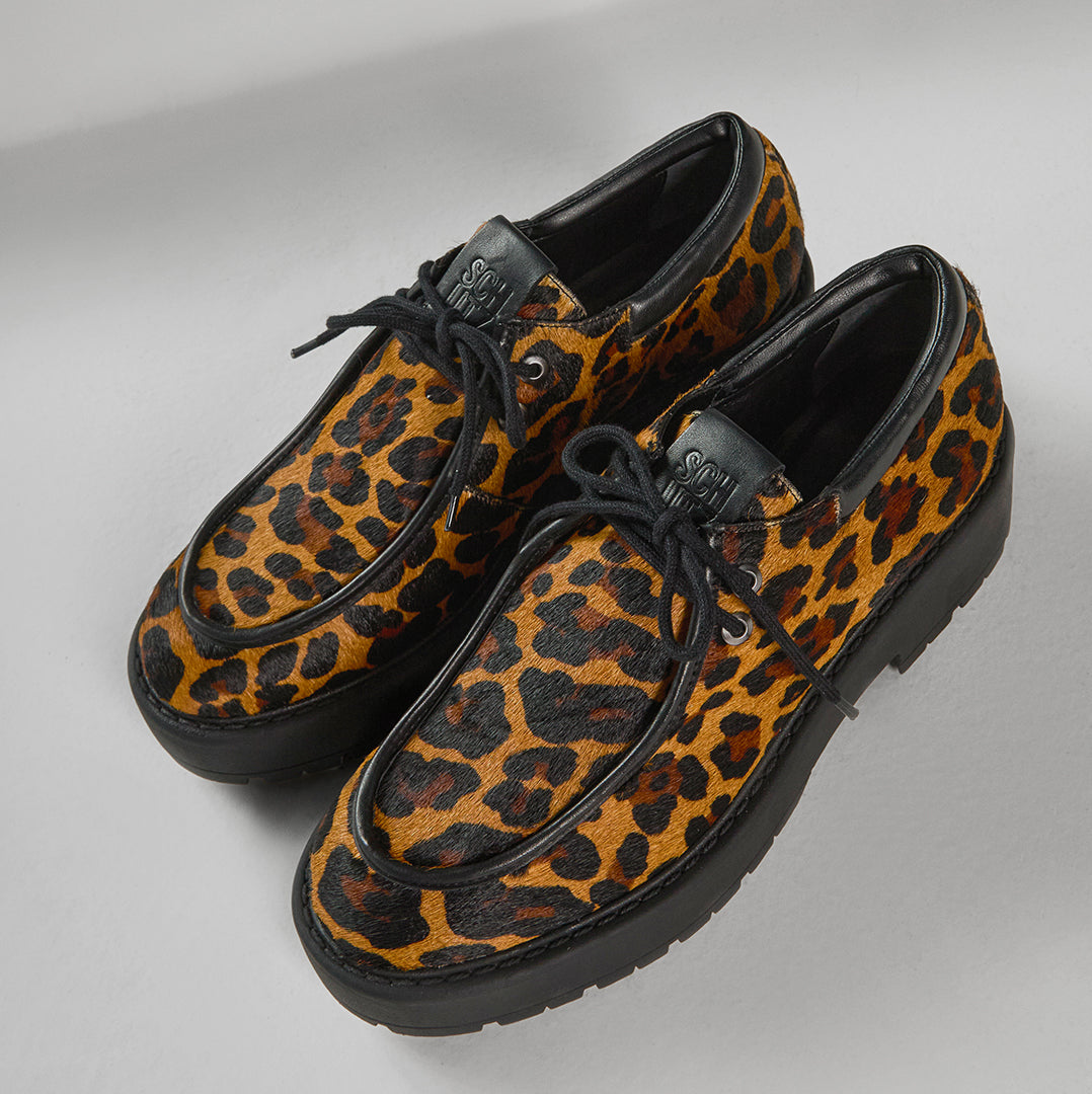 A pair of stylish loafers in a bold leopard print, featuring black laces and a thick black sole. The brand "SCHUTZ" is visible on the tongue.