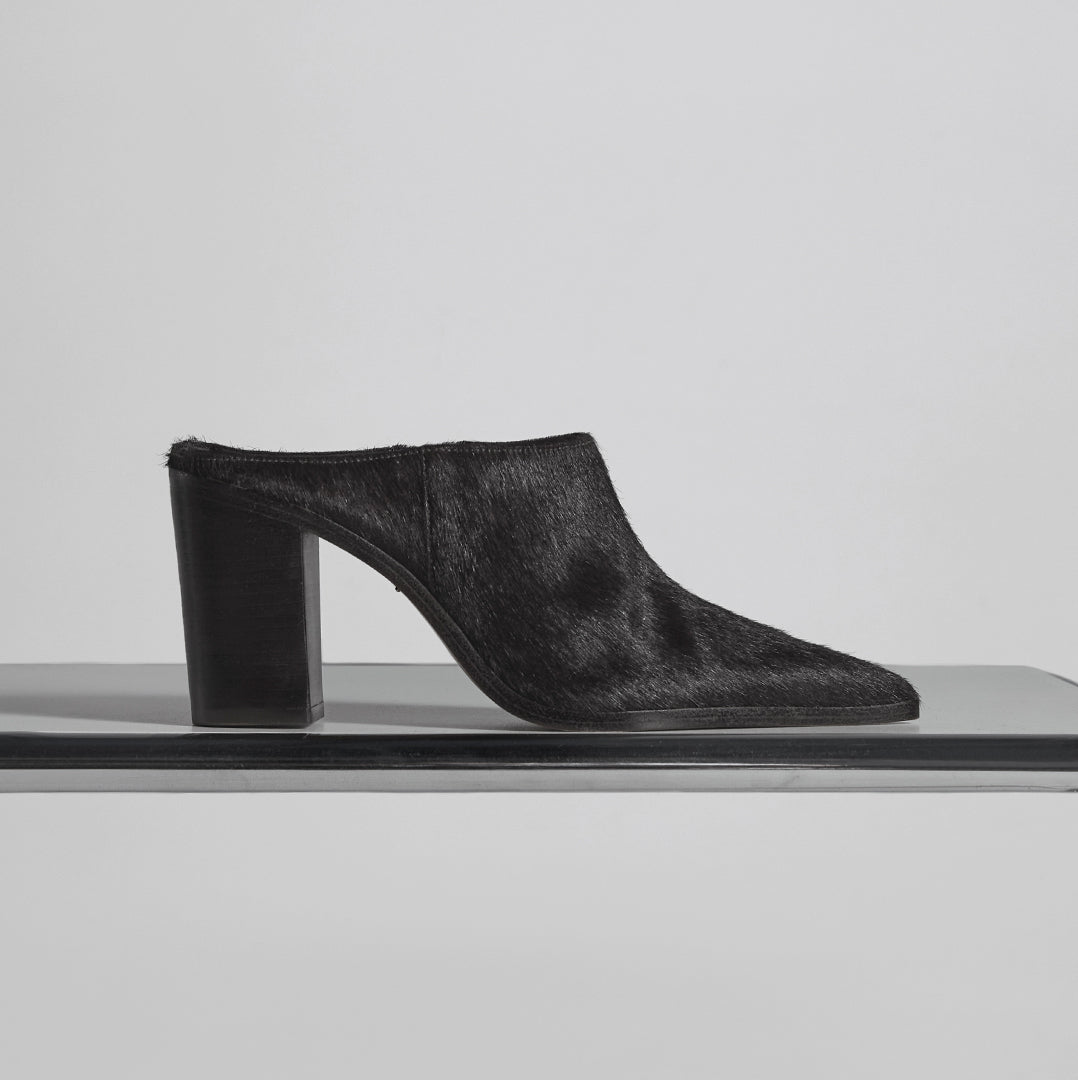 A single black mule shoe with a pointed toe and a chunky block heel, featuring a textured fur upper. The shoe is displayed in profile on a shelf.