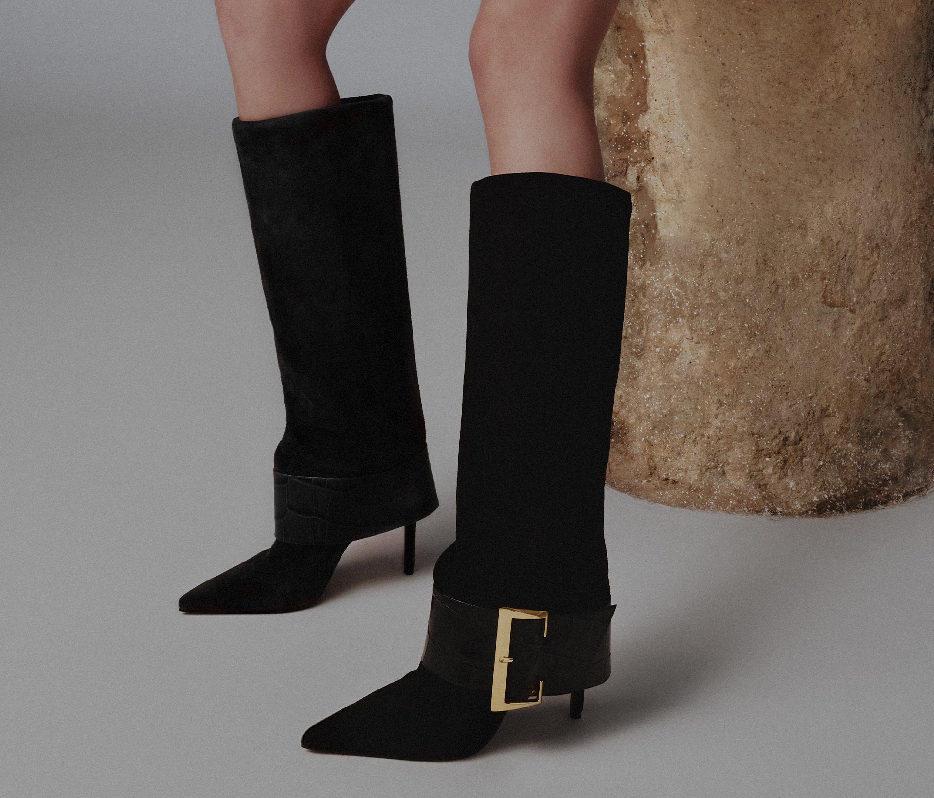 A pair of black suede boots with a sleek, pointed toe and a thick ankle strap with a gold buckle, representing a modern trend.