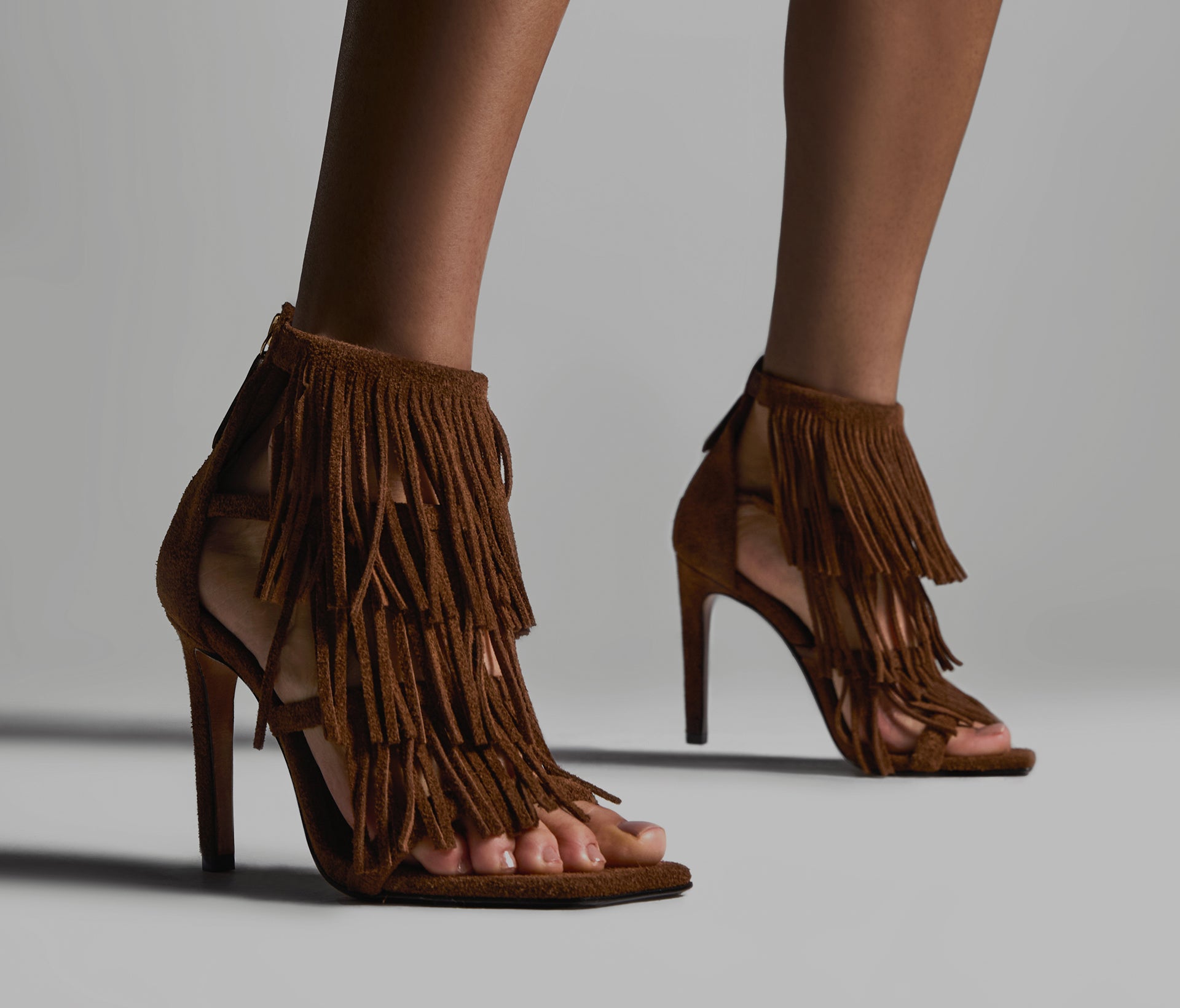 Close-up of a pair of brown suede heeled sandals featuring multiple layers of long, flowing fringe.