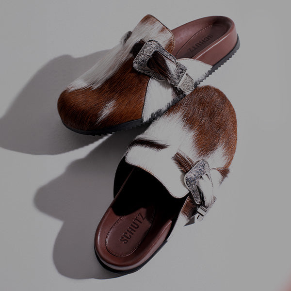 Overhead shot of two slip-on mules or clogs featuring brown and white hair calf material, a round toe, and an ornate silver Western-style buckle decoration.
