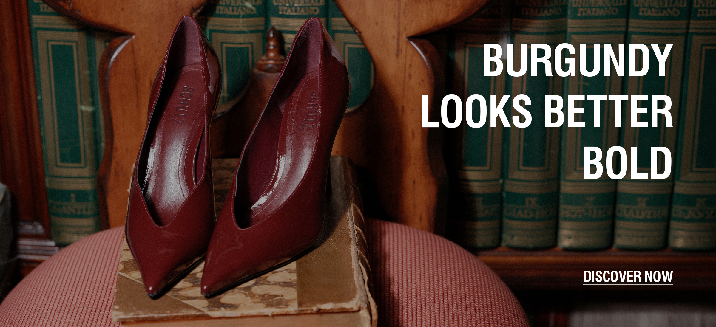 A pair of pointed-toe high heels in a deep burgundy leather, displayed on a vintage book atop a pink upholstered chair, with a background of bookshelves and bold text overlay reading 'BURGUNDY LOOKS BETTER BOLD' and 'DISCOVER NOW'.


