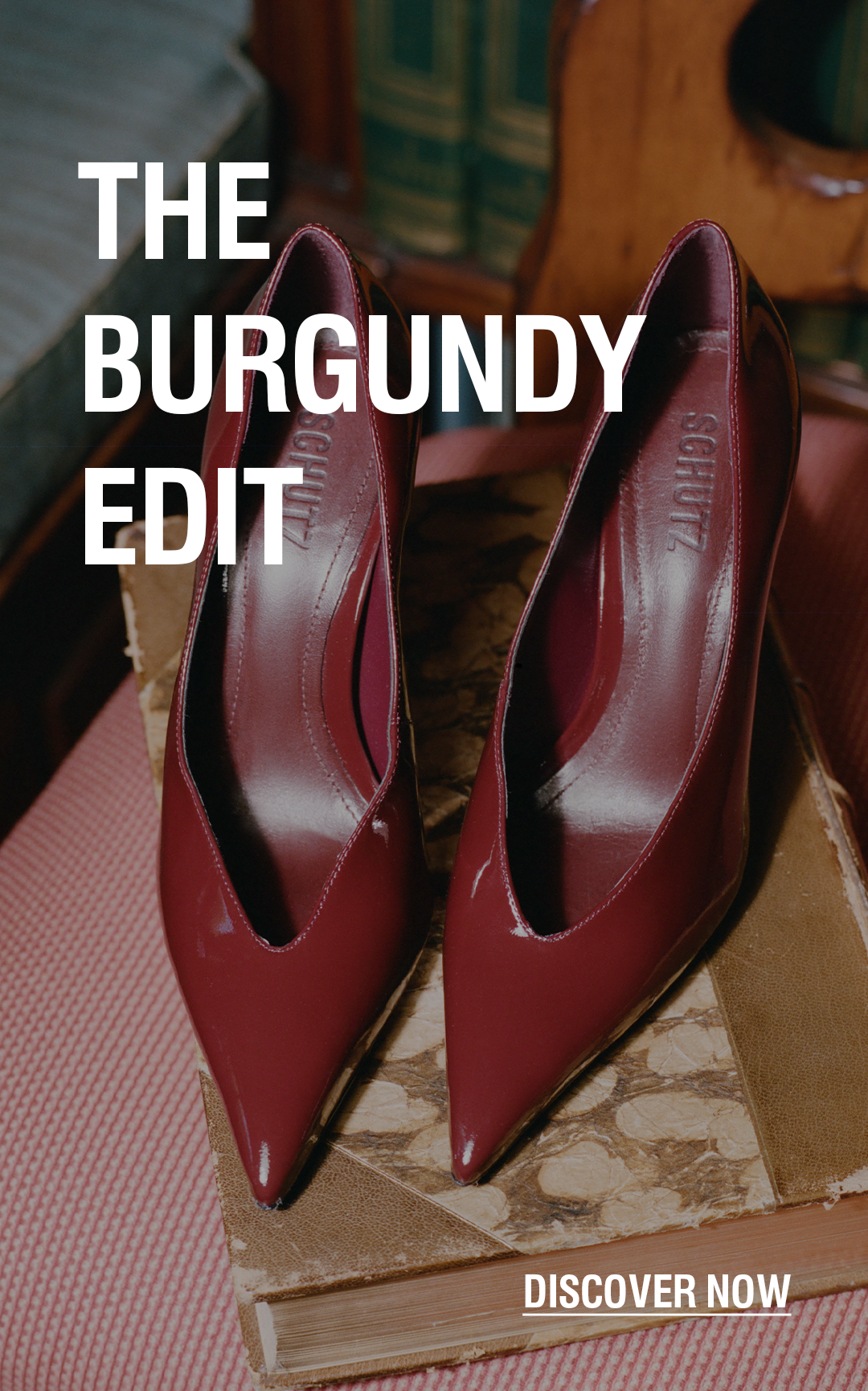 A pair of pointed-toe high heels in a deep burgundy leather, displayed on a vintage book atop a pink upholstered chair, with a background of bookshelves and bold text overlay reading 'BURGUNDY LOOKS BETTER BOLD' and 'DISCOVER NOW'.