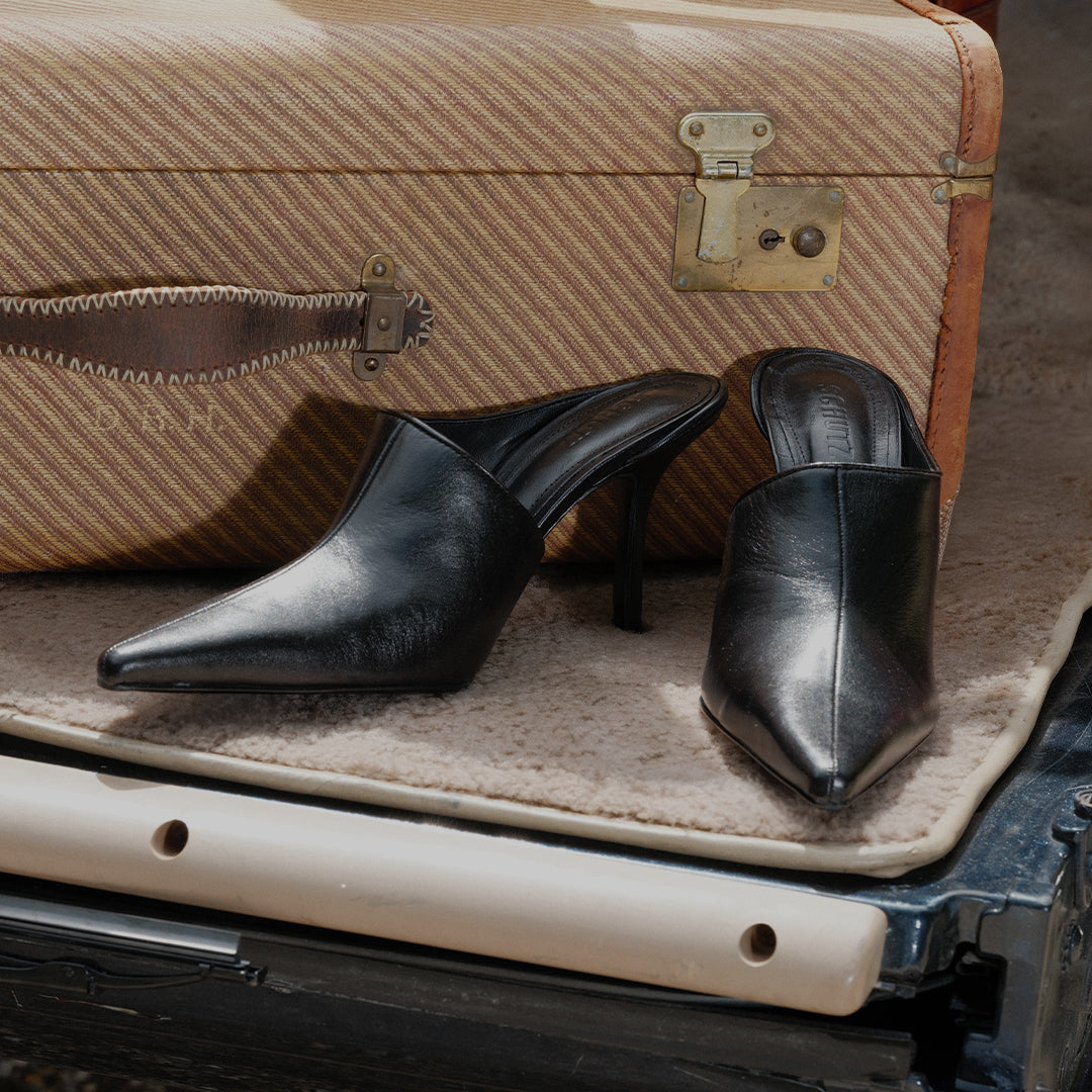 A pointed-toe mule pump with an asymmetrical upper in black leather, placed in front of a vintage suitcase inside a car.