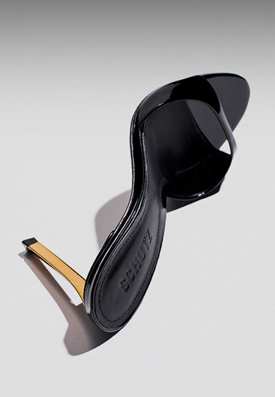 Elegant black patent mule sandal featuring a glossy finish, open toe, and a thin metallic gold heel, placed on a light neutral background.