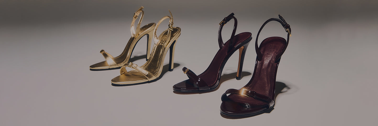 Pair of glossy dark stiletto sandals with thin straps and gold buckles, placed on a soft-lit background with the text ‘STILETTOS SANDALS.