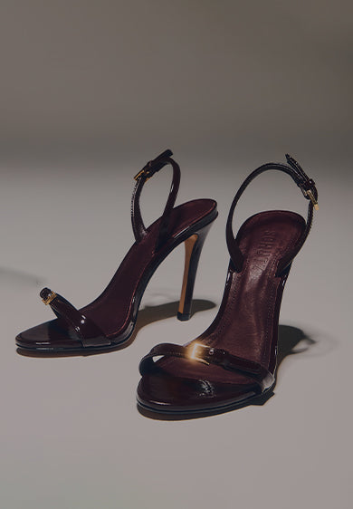 Pair of glossy dark stiletto sandals with thin straps and gold buckles, placed on a soft-lit background with the text ‘STILETTOS SANDALS.