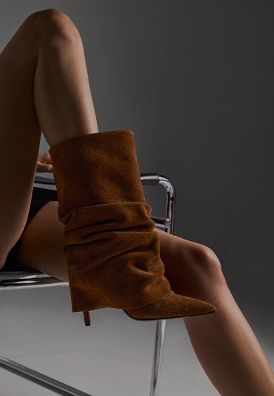 Close-up of a person wearing slouchy brown suede heeled boots while seated on a modern chair, with the text ‘BEST SELLERS.