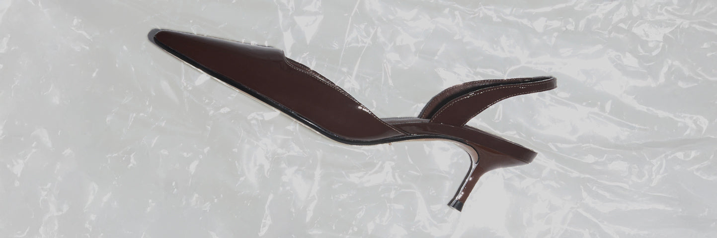 Brown pointed-toe kitten heel shoe with a glossy finish and low slender heel, set against a light textured backdrop.