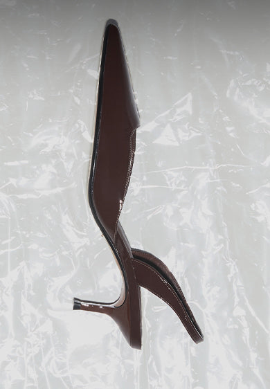 Brown pointed-toe kitten heel shoe with a glossy finish and low slender heel, set against a light textured backdrop.