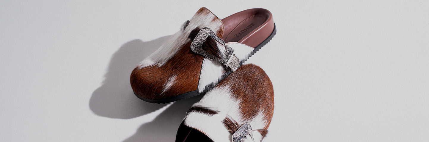 Pair of slip-on flat mules in brown and white cowhide-effect leather with decorative silver buckles, combining rustic texture with modern casual style.