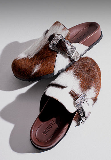 Pair of slip-on flat mules in brown and white cowhide-effect leather with decorative silver buckles, combining rustic texture with modern casual style.