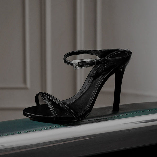Black high-heel sandals with a sleek minimalist design, featuring thin straps and a stiletto heel. The pair is positioned on a reflective teal surface with soft indoor lighting highlighting the glossy finish.