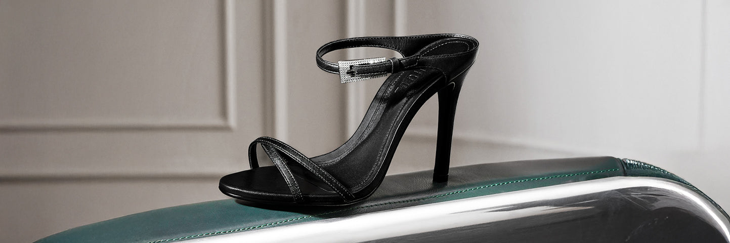 Black high-heeled sandal with ankle strap displayed on a green and silver surface, with the text ‘WOMEN’S SHOES’ on the left.