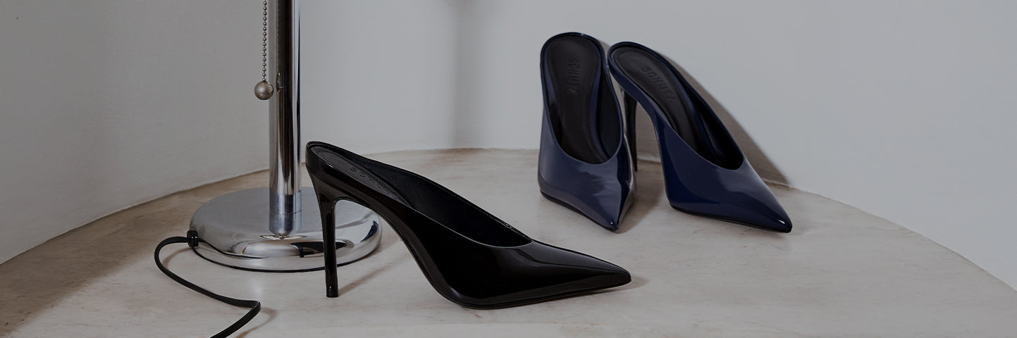 Two pairs of high-heeled pointed-toe pumps displayed beside a chrome floor lamp, one in glossy black and the other in glossy navy.