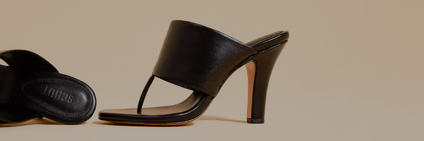 Women’s black leather thong sandals with a sleek mid-height stiletto heel, photographed in a minimalist studio setting with soft neutral tones.