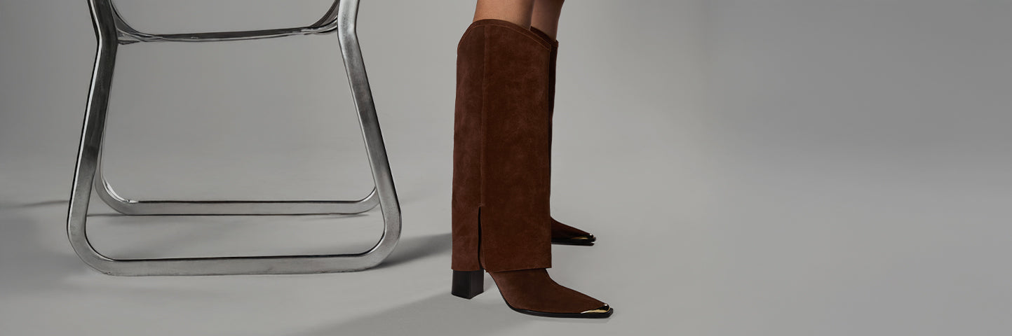 Close-up of brown suede knee-high boots with gold-tipped toes, styled next to a metal chair.