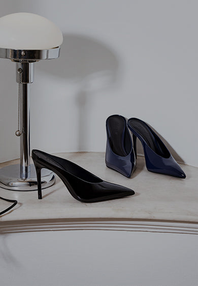 Two pairs of high-heeled pointed-toe pumps displayed beside a chrome floor lamp, one in glossy black and the other in glossy navy.