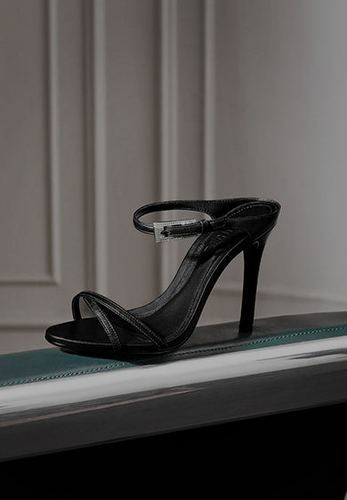 Black high-heeled sandal with ankle strap displayed on a green and silver surface, with the text ‘WOMEN’S SHOES’ on the left.