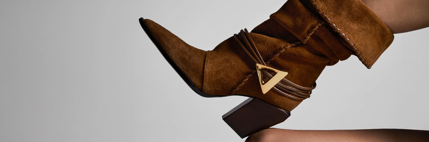 A stylish suede brown ankle boot with a pointed toe and chunky heel, decorated with a gold triangular buckle detail. The boot is shown against a gray background with the text “BROWN SHOES