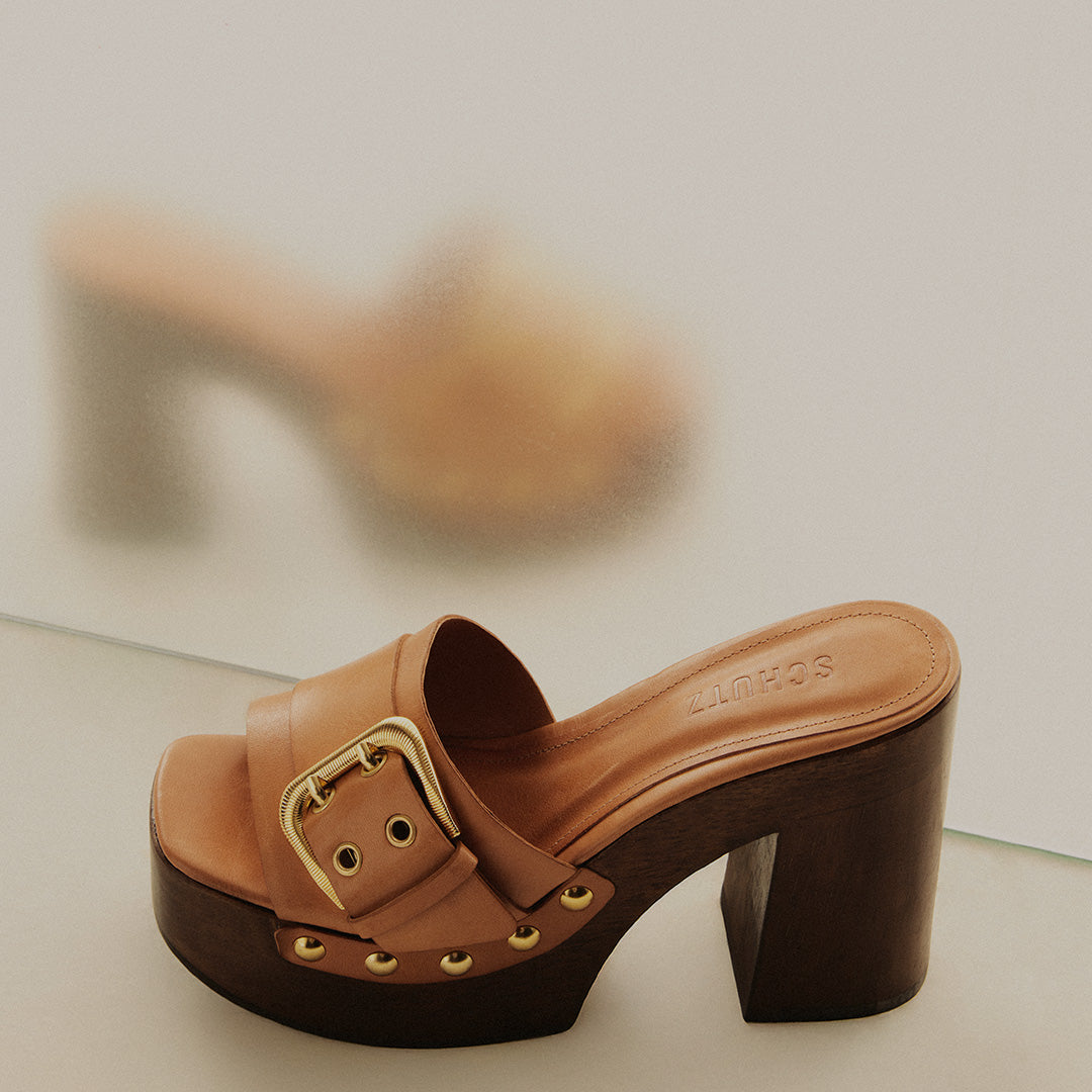 A brown leather wooden-heel mule sandal with a thick platform sole and buckle detail, displayed against a neutral background. Text above reads “Wooden Heels” and “Shop New In.”