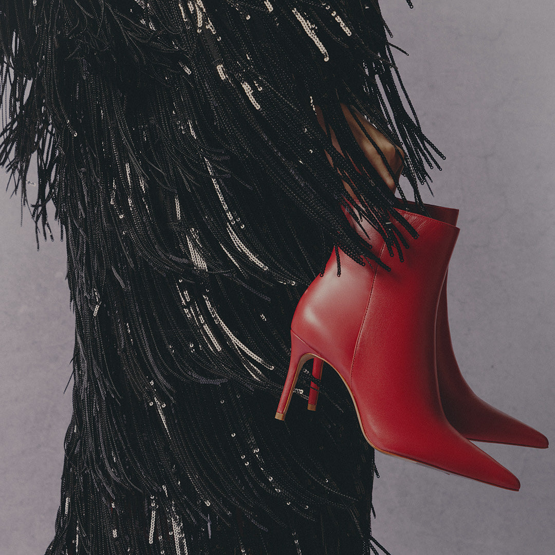 Close-up of a model in a black fringed and sequined outfit holding red pointed-toe high-heeled ankle boots; overlay text reads “DATE NIGHT STYLES – SHOP BOOTS."