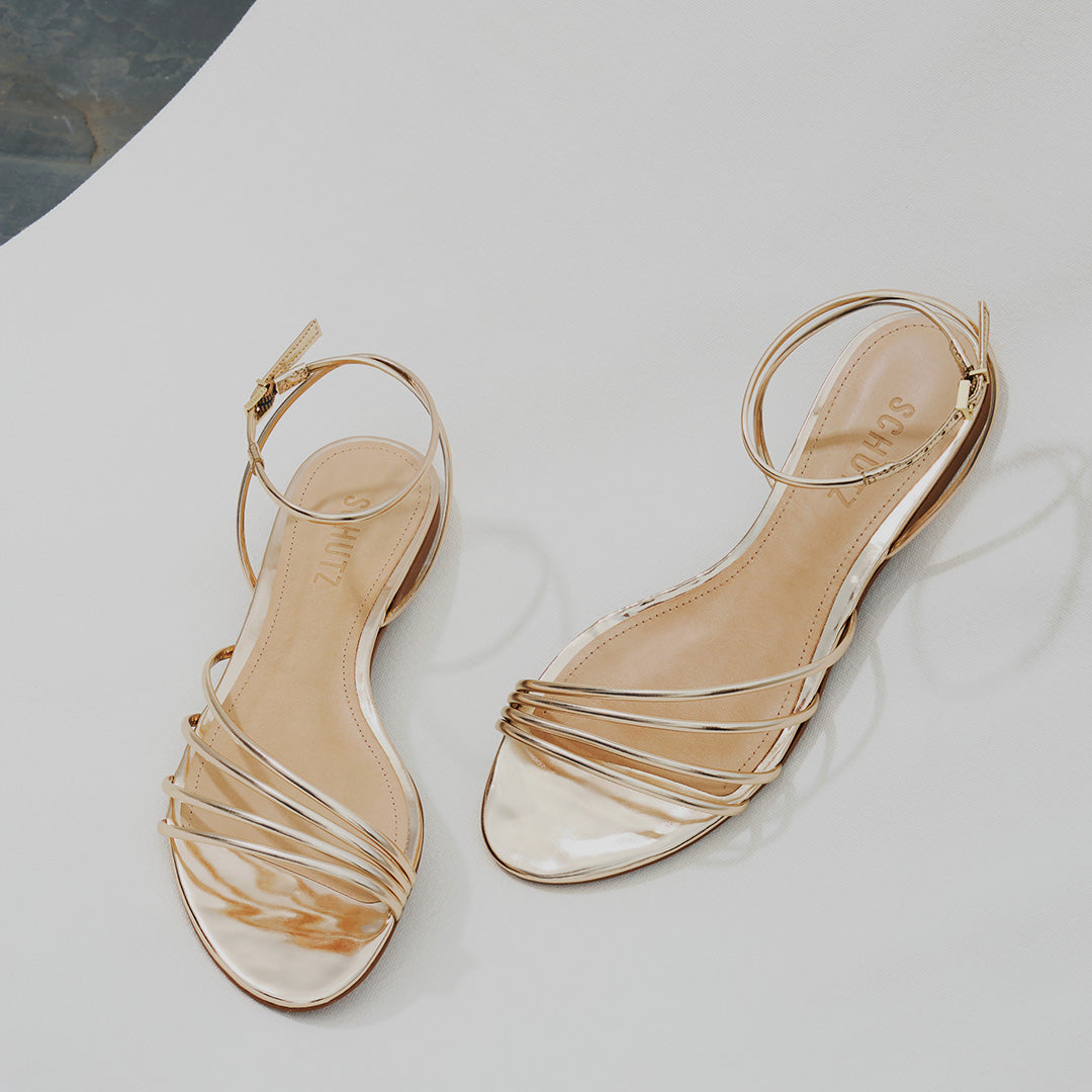 Two elegant, shiny gold sandals with thin straps are placed on a smooth, light surface. Each features an ankle strap and a reflective finish.