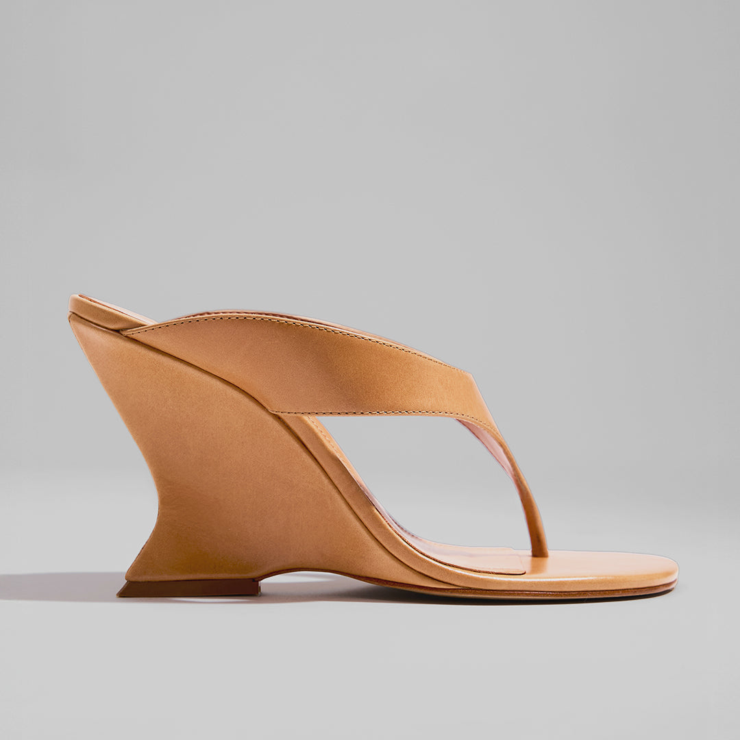 Tan wedge sandal with a modern, sculpted heel design. Features a wide strap and thong style on a plain gray background, conveying elegance.