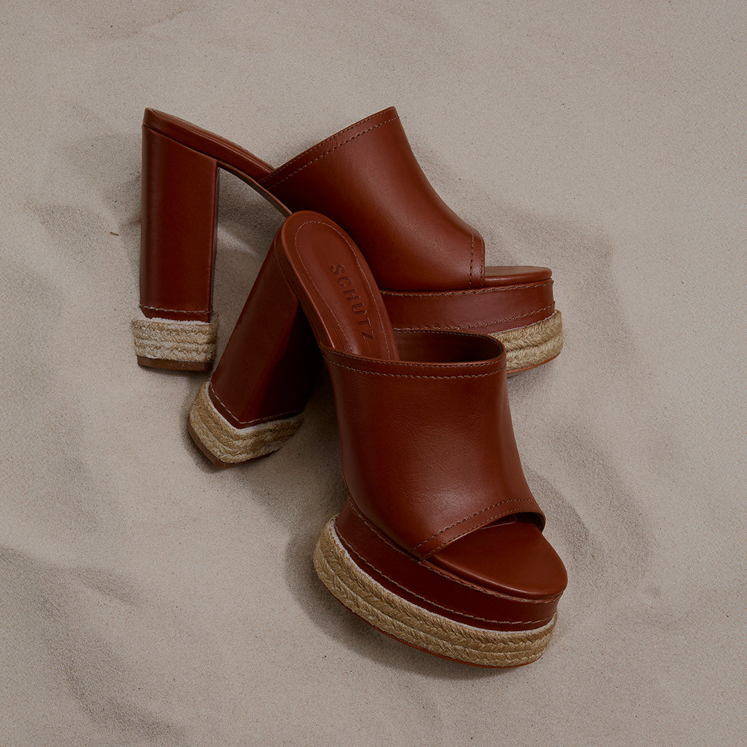 Brown leather platform sandals with high block heels, featuring rope detailing, placed on light beige sand, creating a summery, casual vibe. Tap to Shop Platforms.