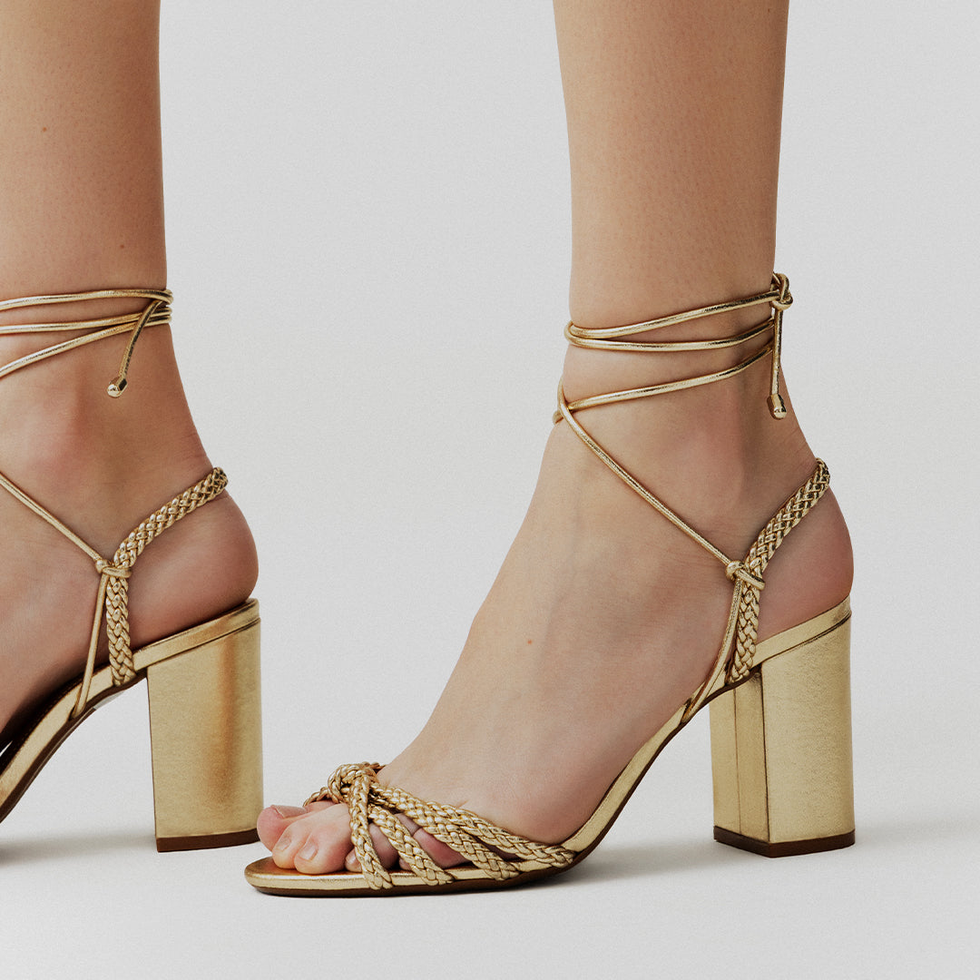 Gold strappy high-heel sandals with braided straps and ankle ties on a woman's feet, against a neutral background, convey elegance and sophistication.