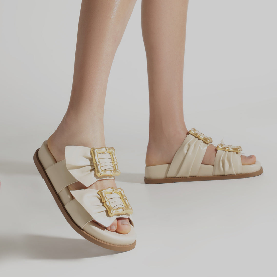 Close-up of a person wearing stylish cream sandals with large gold buckles and bows. The sandals have a flat sole and exude a chic, modern vibe.