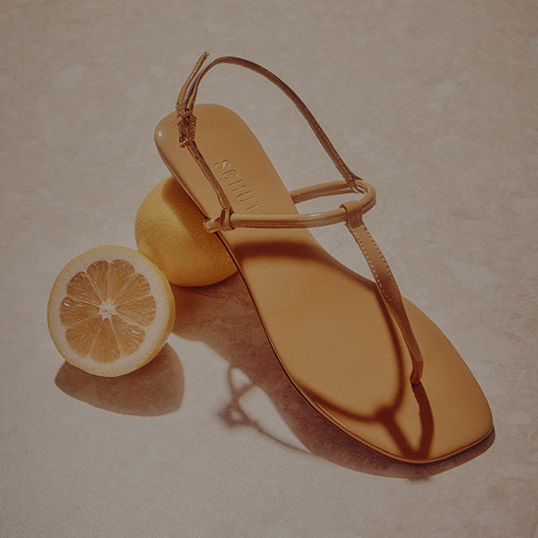 A tan sandal rests on a table, artistically balanced on a whole lemon with a halved lemon beside it. The lighting casts soft shadows, creating a relaxed, summery feel. 