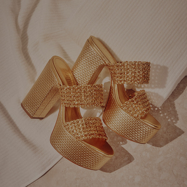Golden platform sandals with woven straps and high block heels are stylishly paired resting on a textured cream fabric, exuding elegance and glamour.