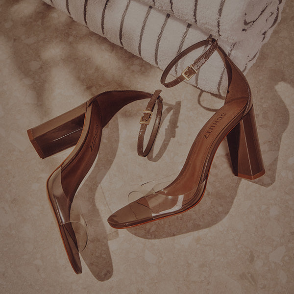 Brown block heel sandals with clear straps andgold buckles rest on a light stone floor near a folded striped towel, conveyingan elegant and stylish tone.