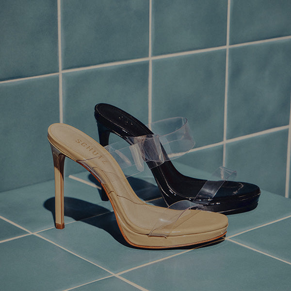 Two high-heeled shoes with clear straps are set on teal tiles. One shoe has a beige sole, the other black, evoking a modern, minimalist style.