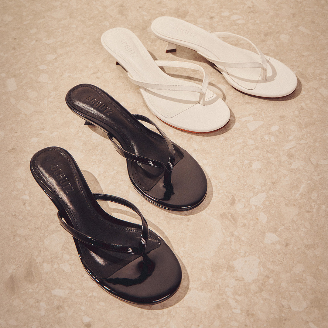 Two pairs of heeled sandals on a textured beige floor. One pair is glossy black and the other is matte white, elegantly arranged for a chic display.