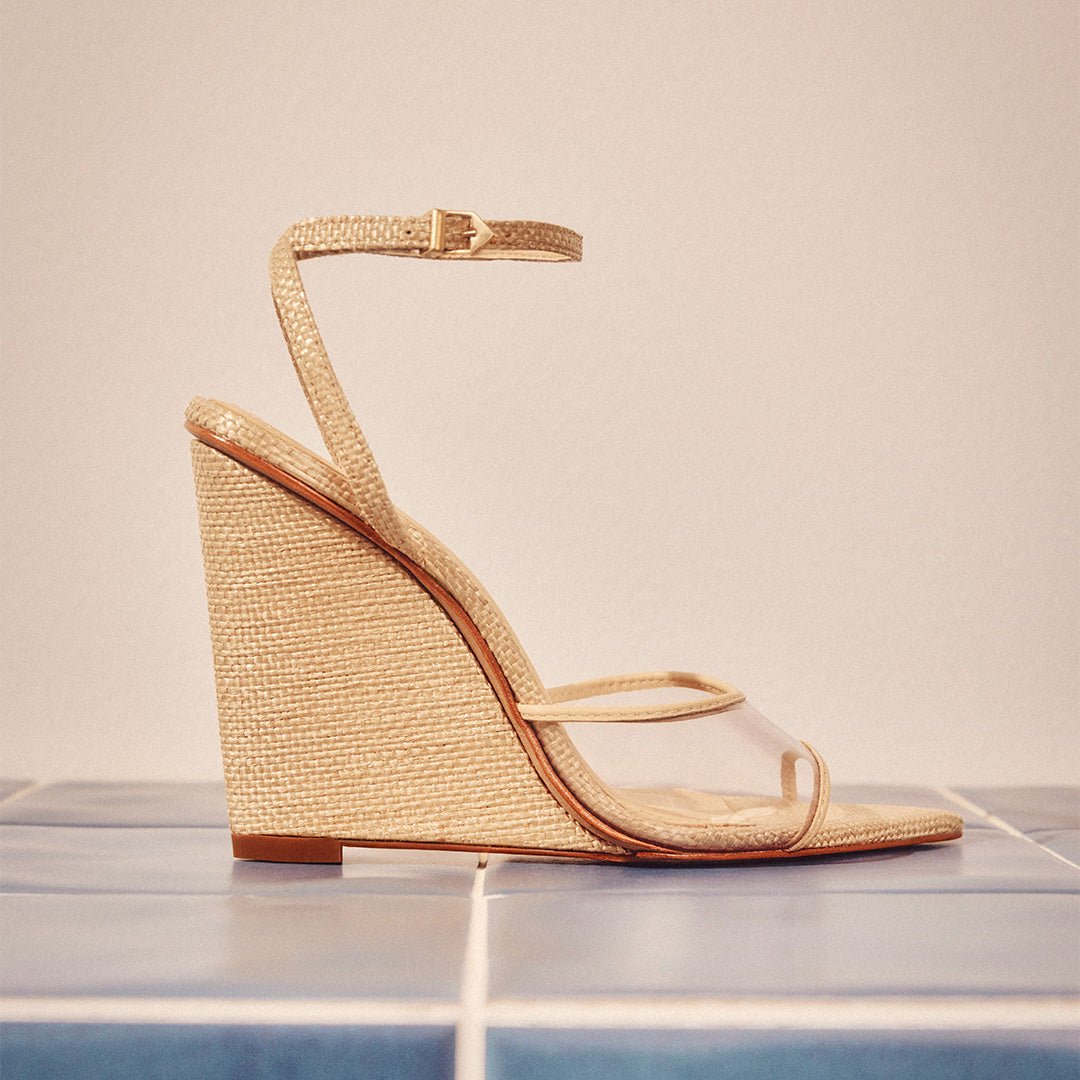 Beige wedge sandal with a transparent strap and ankle buckle on a blue-tiled surface. The minimalistic design gives a summery, elegant vibe.