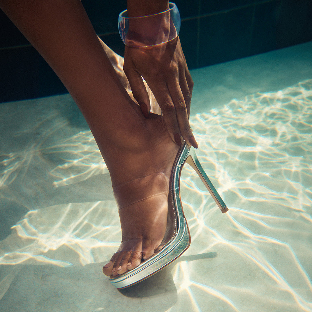 A hand adjusts a clear high-heeled shoe on a foot submerged in a sunlit pool. Water reflections create a serene, summery vibe.
