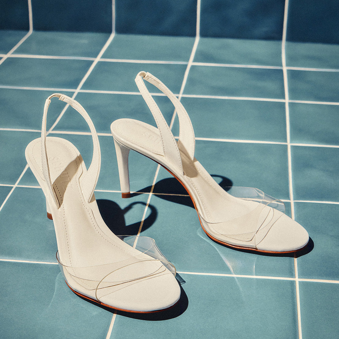White slingback heels with clear straps are placed on sunlit blue tiles. The scene conveys elegance and a summery, sophisticated tone.