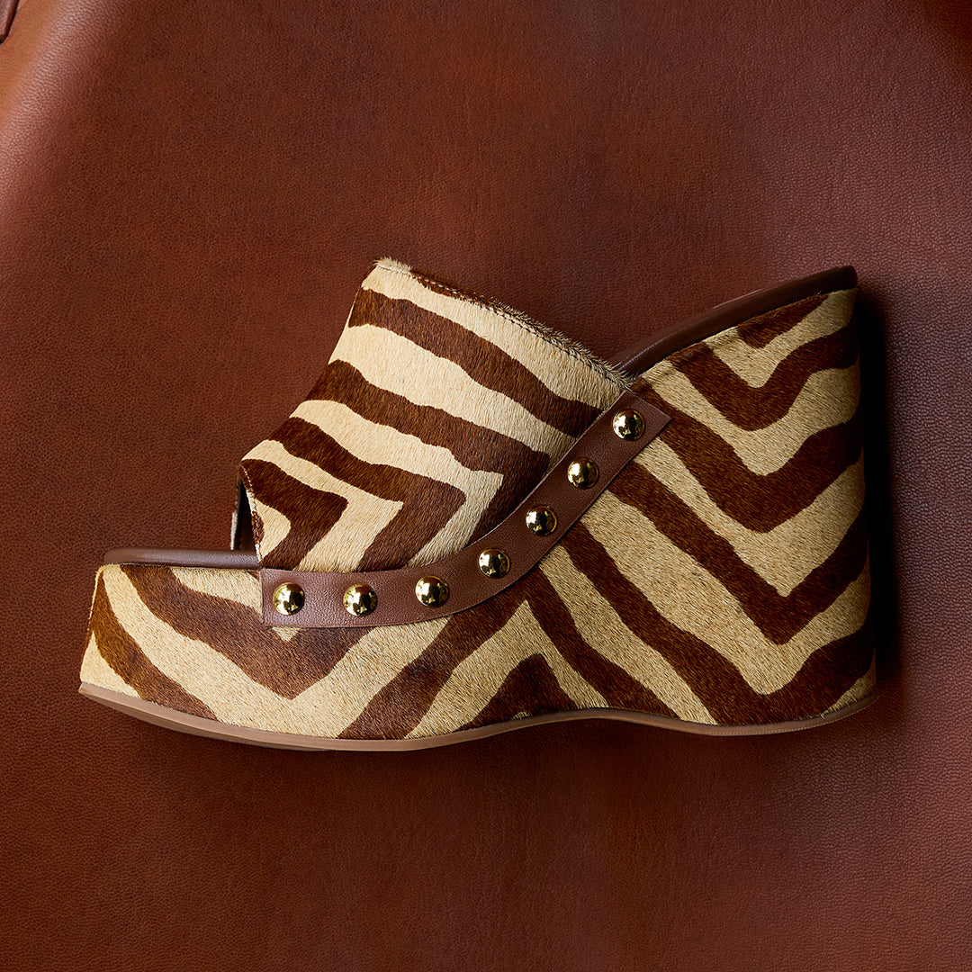 A stylish platform clog shoe with a zebra pattern and golden studs, set against a rich brown leather background, exuding a bold, trendy vibe.