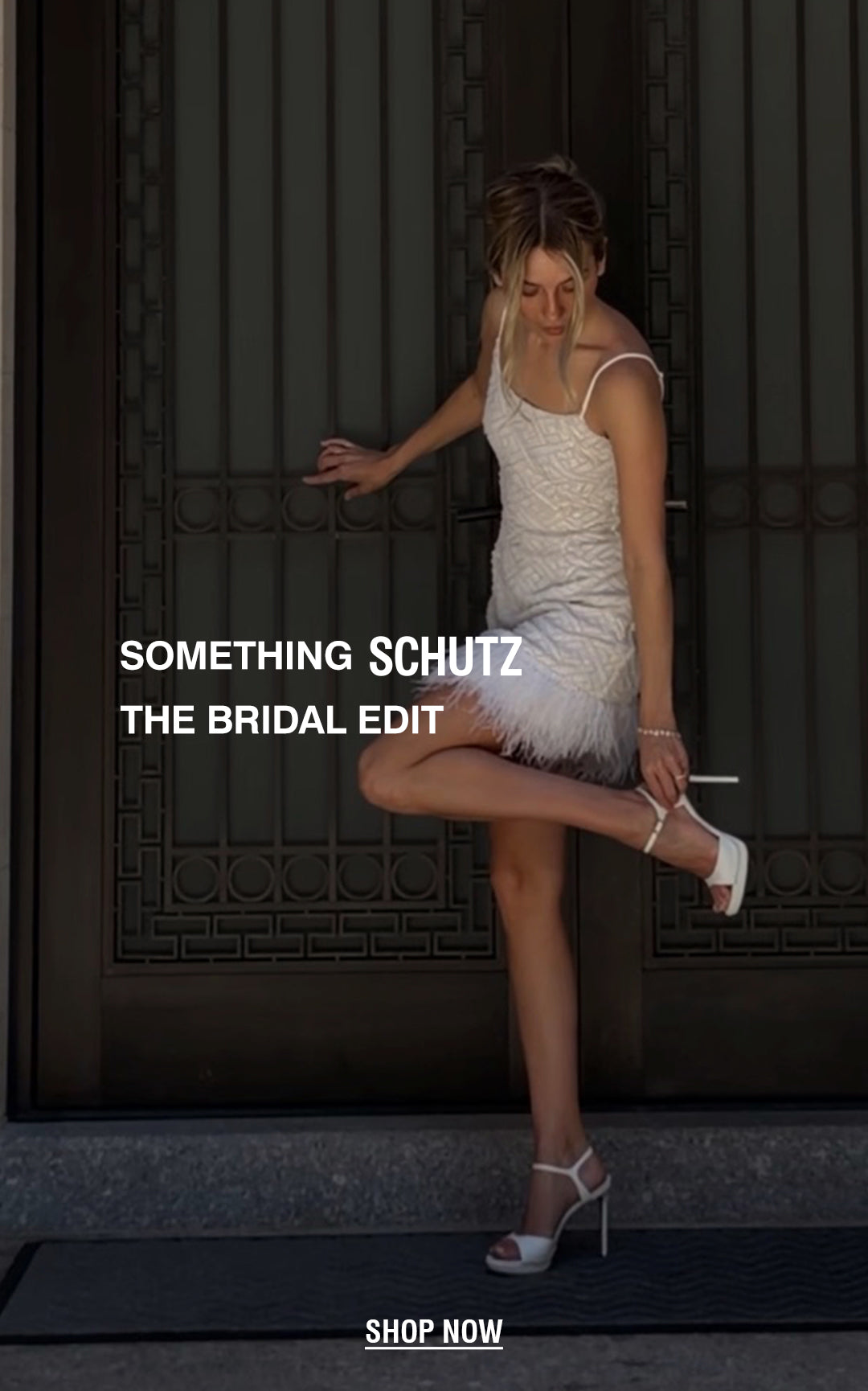 Alt text: Young woman in white feathered dress adjusts her white high heels beside a metal door. Text reads "Something Schutz" and "The Bridal Edit." Tone is elegant and stylish.