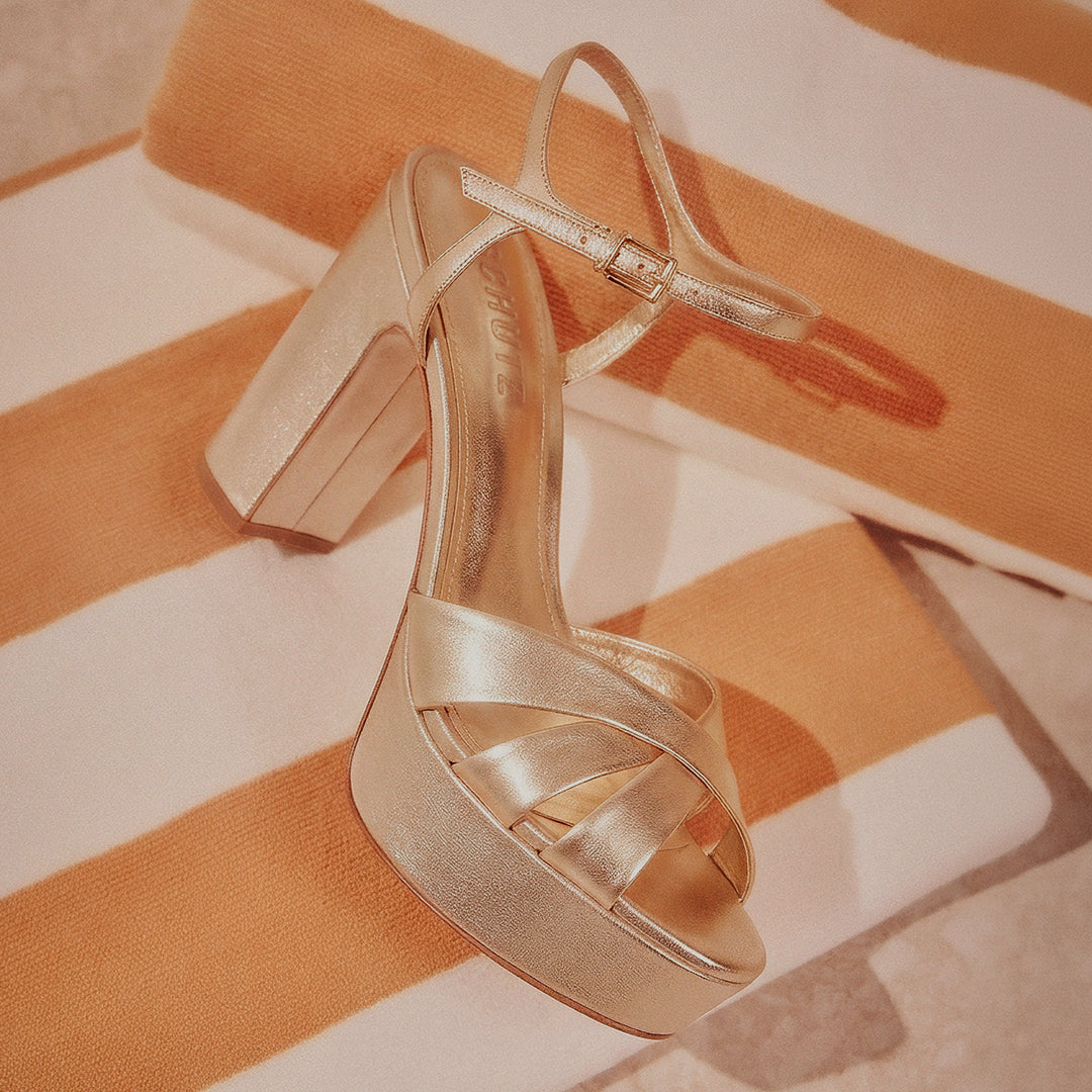 A gold high-heeled sandal with an ankle strap is placed on an orange and white striped background, conveying elegance and style.