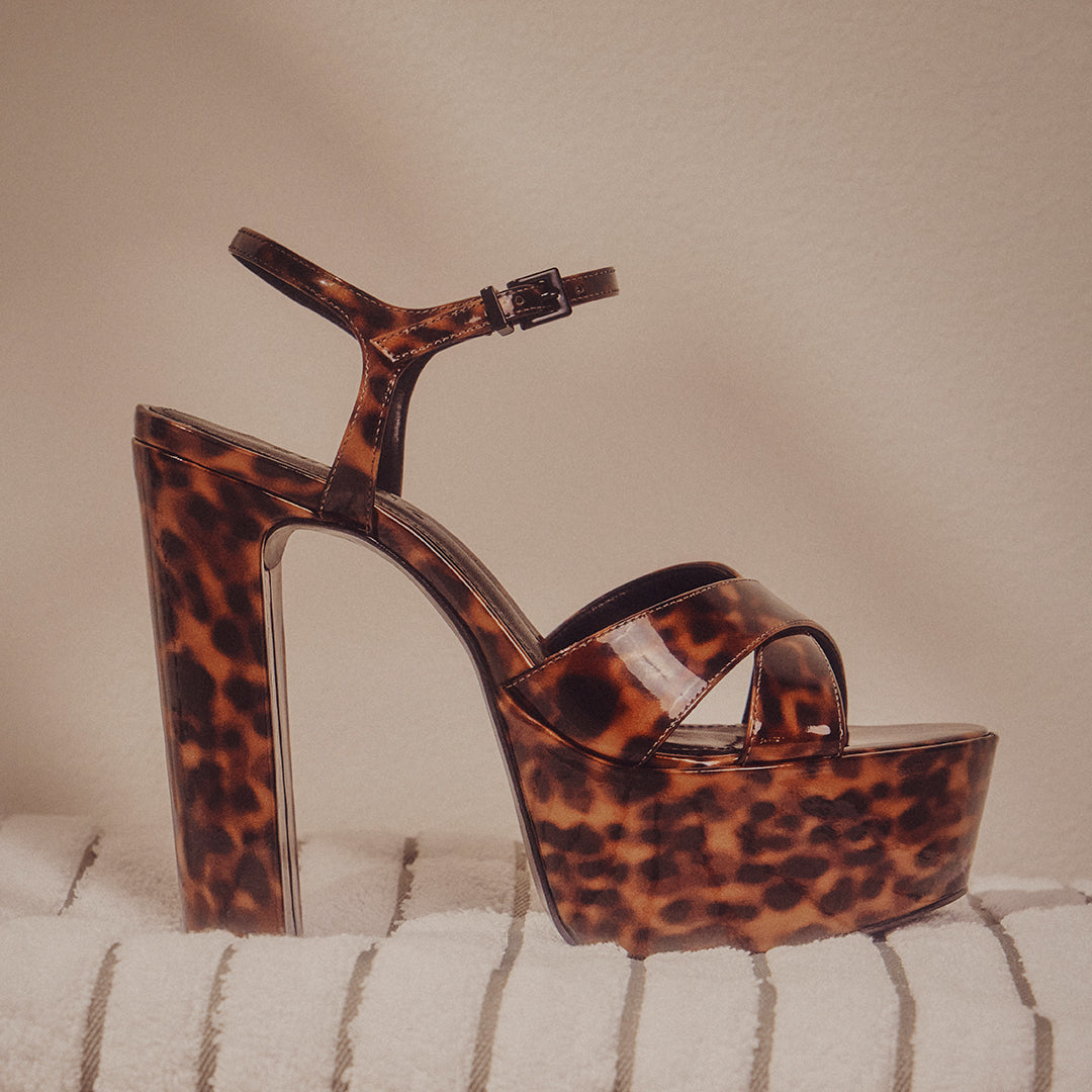 A single leopard print platform heel with an ankle strap sits on textured fabric. The shoe exudes a bold, stylish, and fashion-forward vibe.