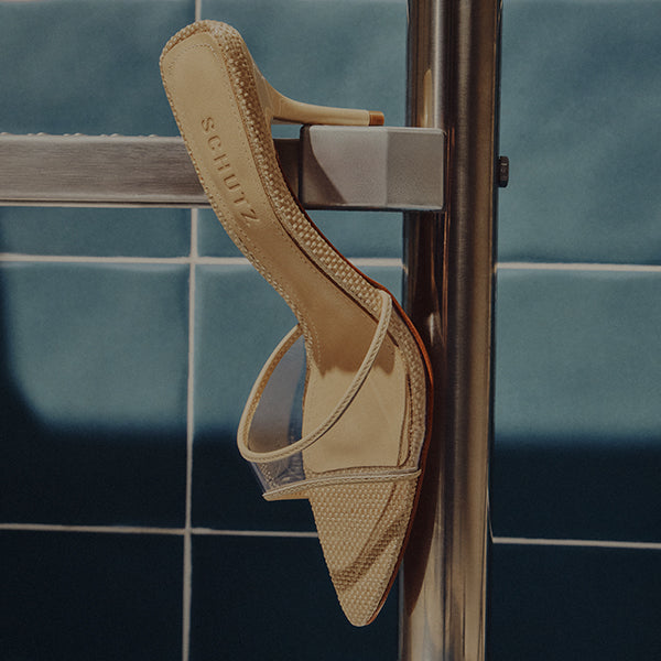 A beige high-heeled sandal with a translucent strap hangs on a metal bar against a blue tiled wall, creating a minimalist and stylish vibe.